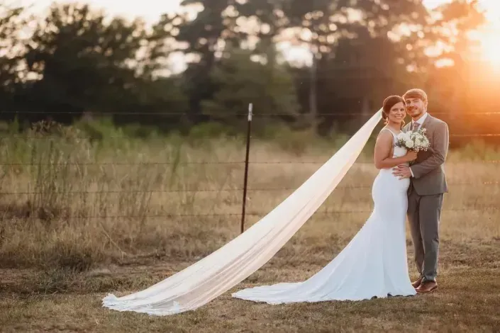 Bride and groom embracing outdoors at sunset, with a long flowing veil trailing behind them