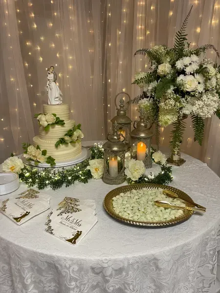 Elegant dessert table with white cake, flowers, candles, and gold serving tray on a draped table.
