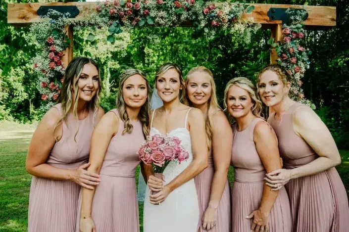 Six women in pink dresses pose with a bride in white holding a bouquet under a floral arch outdoors.