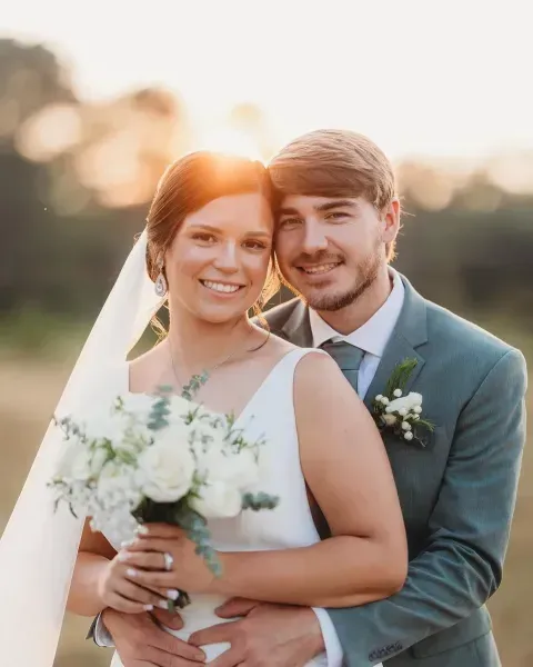 Smiling couple in wedding attire embracing outdoors at sunset, bride holding a white bouquet.