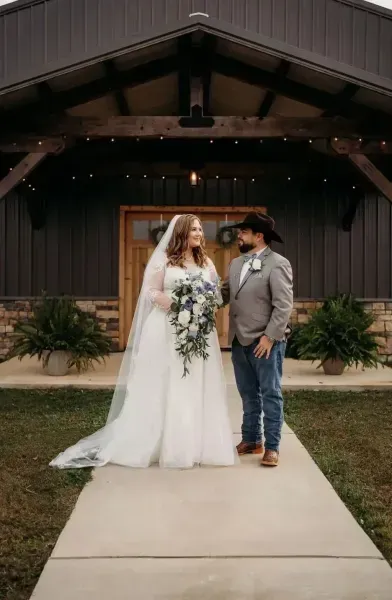 Newlyweds standing outside a rustic venue, bride holding bouquet in a white gown and groom in a hat and jeans