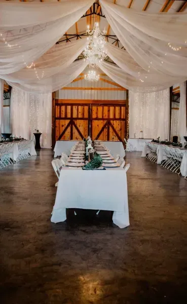 White-draped banquet hall with long tables set for a wedding reception and a chandelier overhead