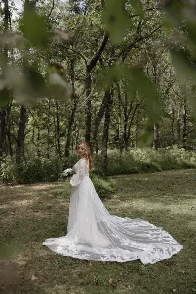 Bride in a long white gown standing in a wooded park, with the dress trailing behind her.