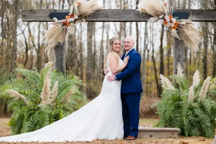 Couple embracing under a rustic floral arch outdoors, with a long white dress and blue suit