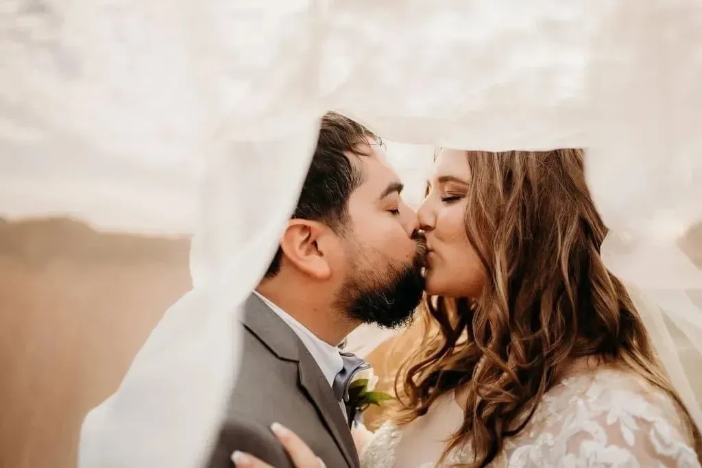 Couple kissing under a veil at an outdoor wedding ceremony
