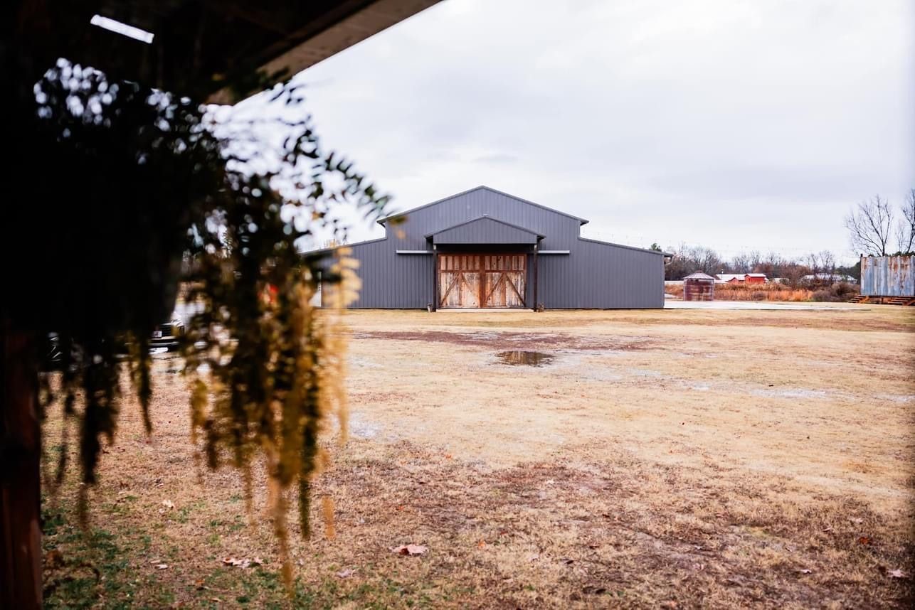 Gray barn-like building in a dry field, seen from a porch with hanging plants in the foreground