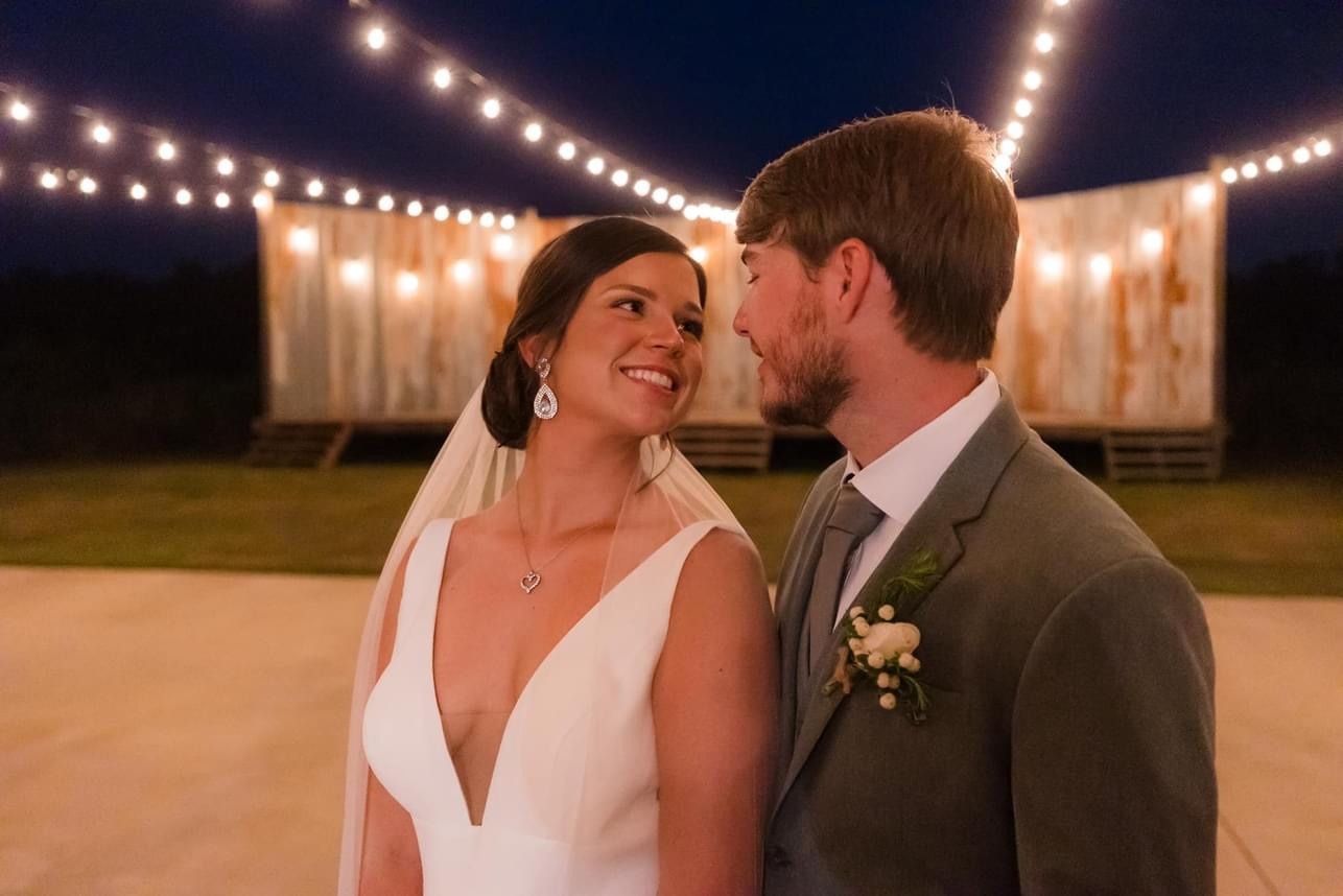 Bride and groom smiling at each other under string lights at an outdoor evening wedding.