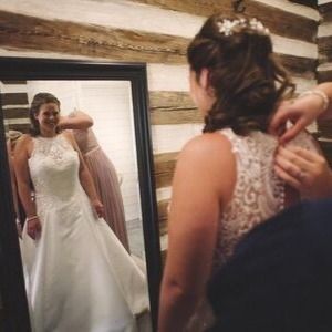 Bride adjusting lace wedding dress in front of a mirror while another person helps with the back straps
