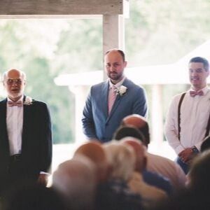 Three men in formalwear standing at a wedding ceremony, facing seated guests.
