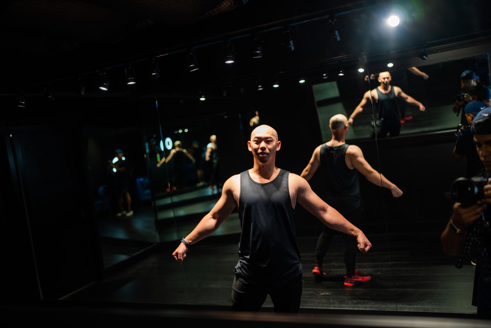 Man in black tank top posing in a mirrored dance studio under dramatic lighting