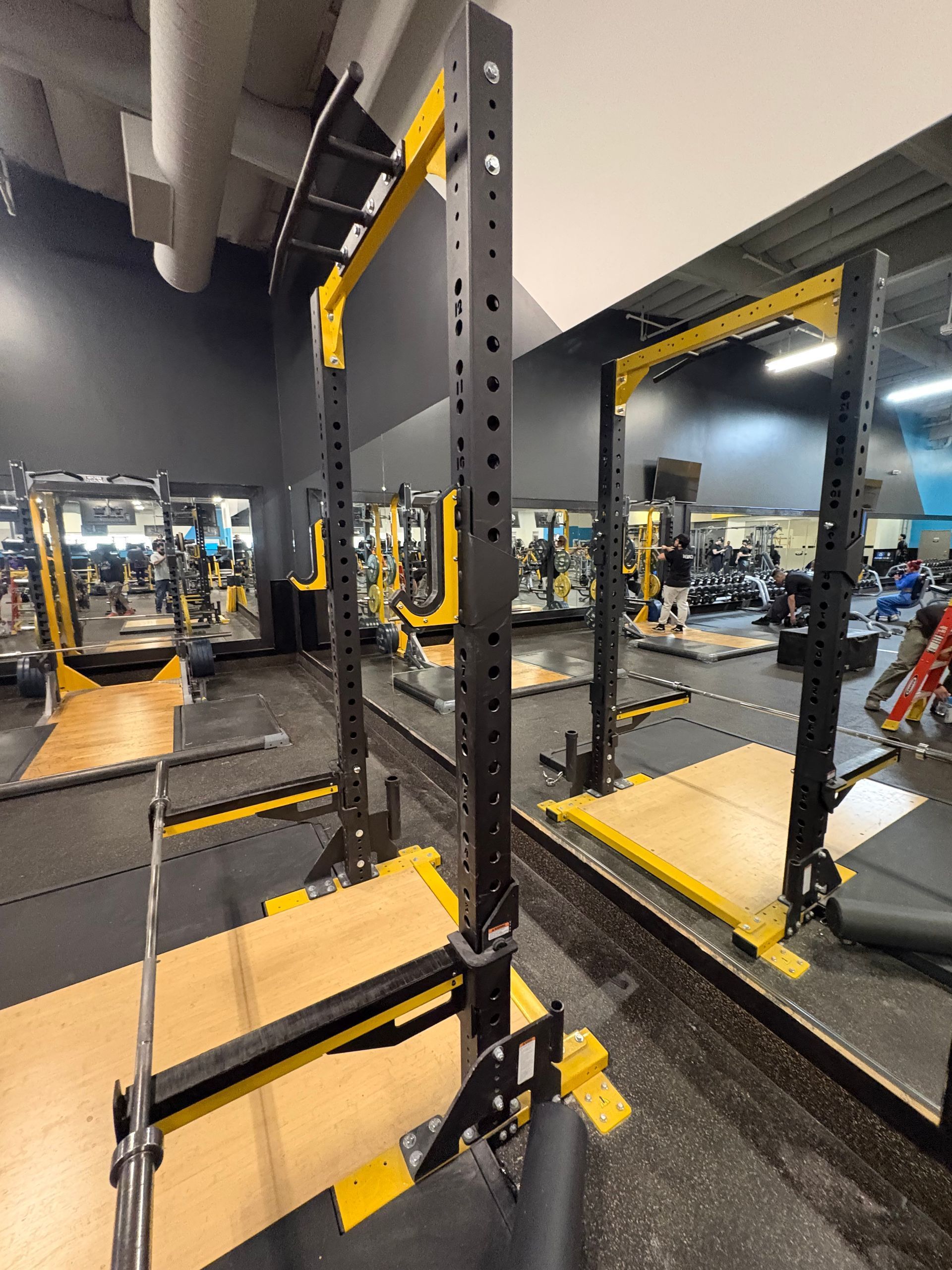 Gym squat rack with barbell, weight plates, and mirrors on a black-and-yellow lifting platform.