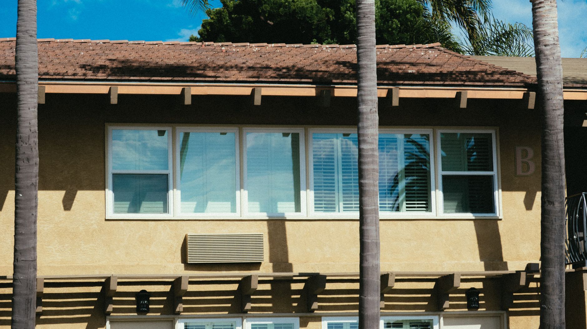 Single-story tan building with windows, benches, and palm trees casting shadows in front