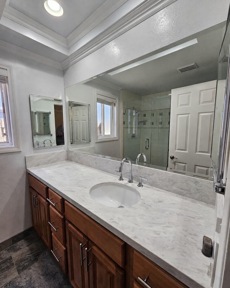 Bright bathroom vanity with large mirror, marble countertop, wooden cabinets, and glass shower in reflection