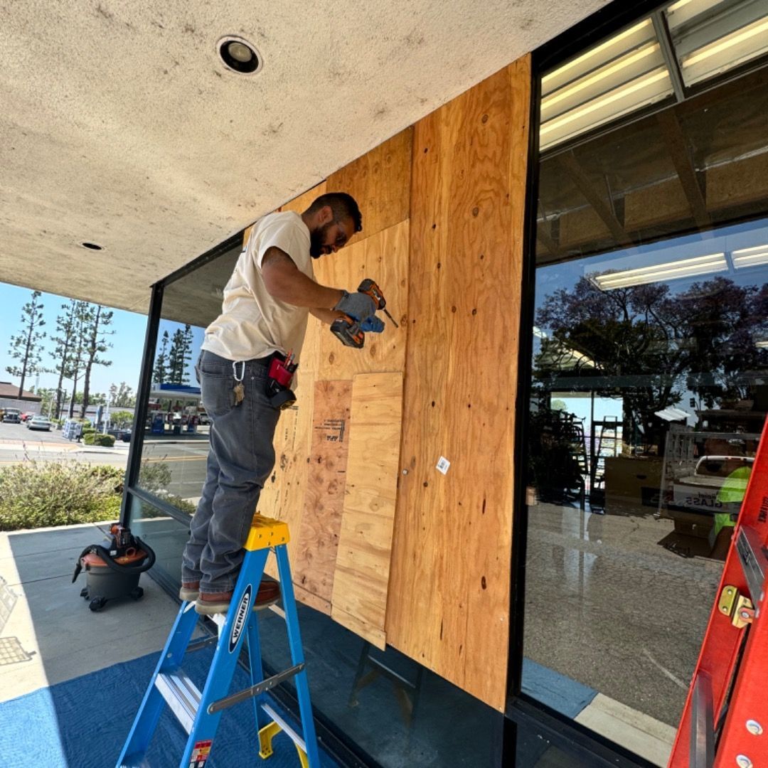 Worker drilling plywood over storefront window under a covered sidewalk.