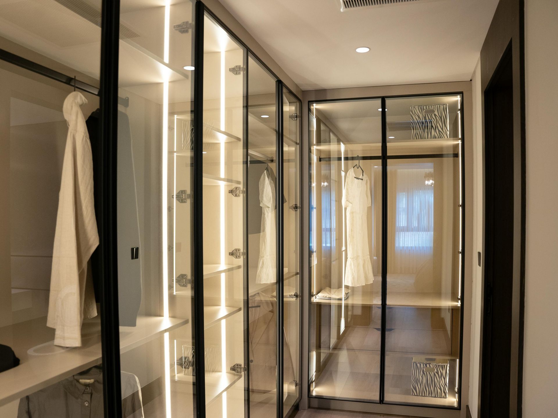 Modern walk-in closet with glass doors, white robes, and shelves in a bright hallway