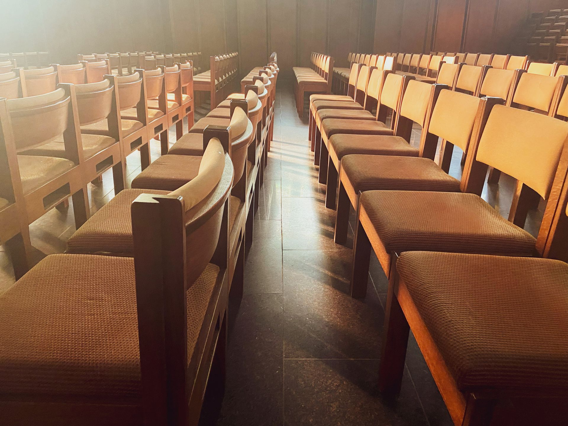 File di sedie di legno vuote in un teatro, immerse nella luce soffusa del palco.