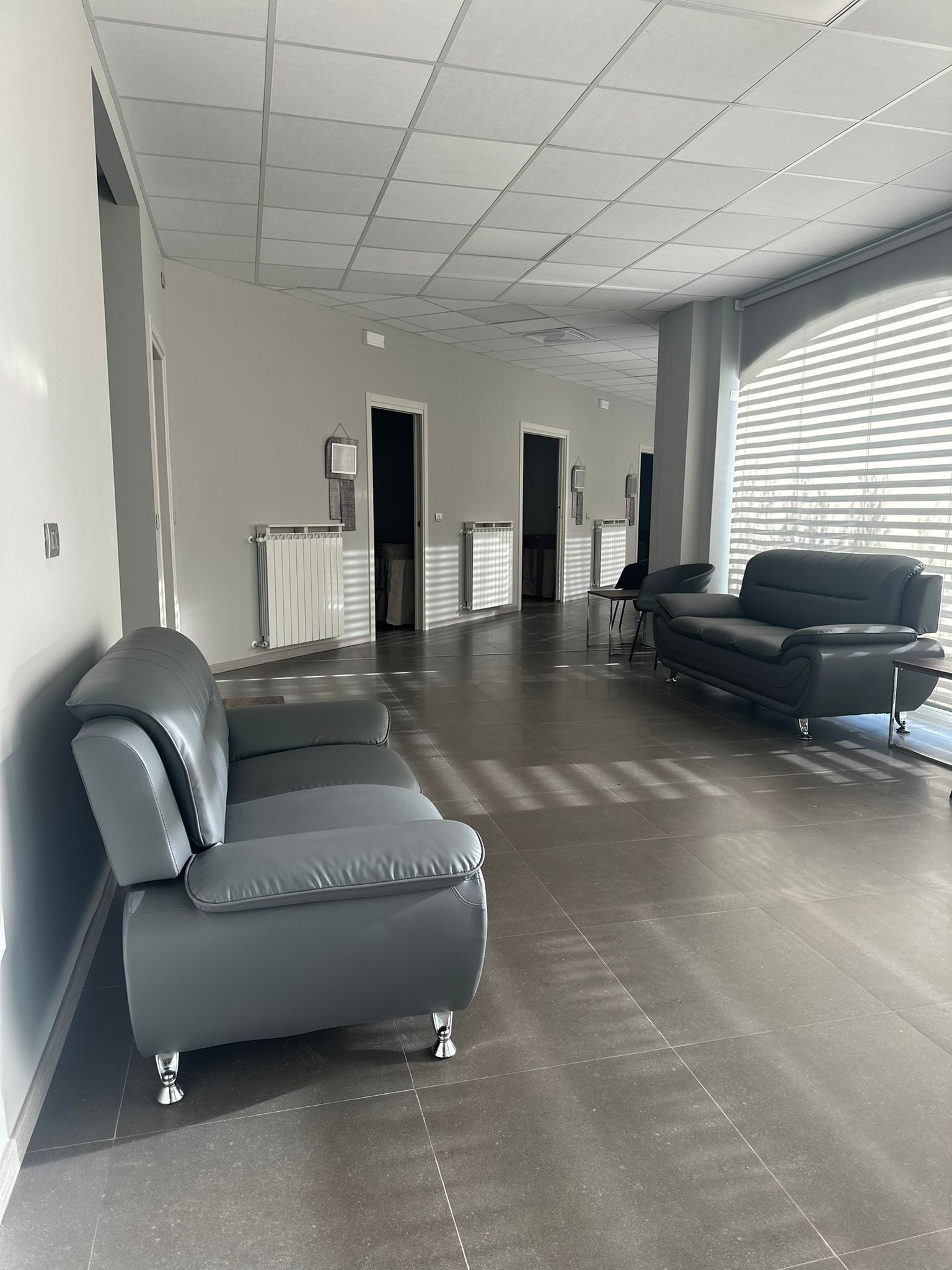 Una lunga sala d'attesa vuota con divani grigi, pavimenti in cemento e finestre con persiane.