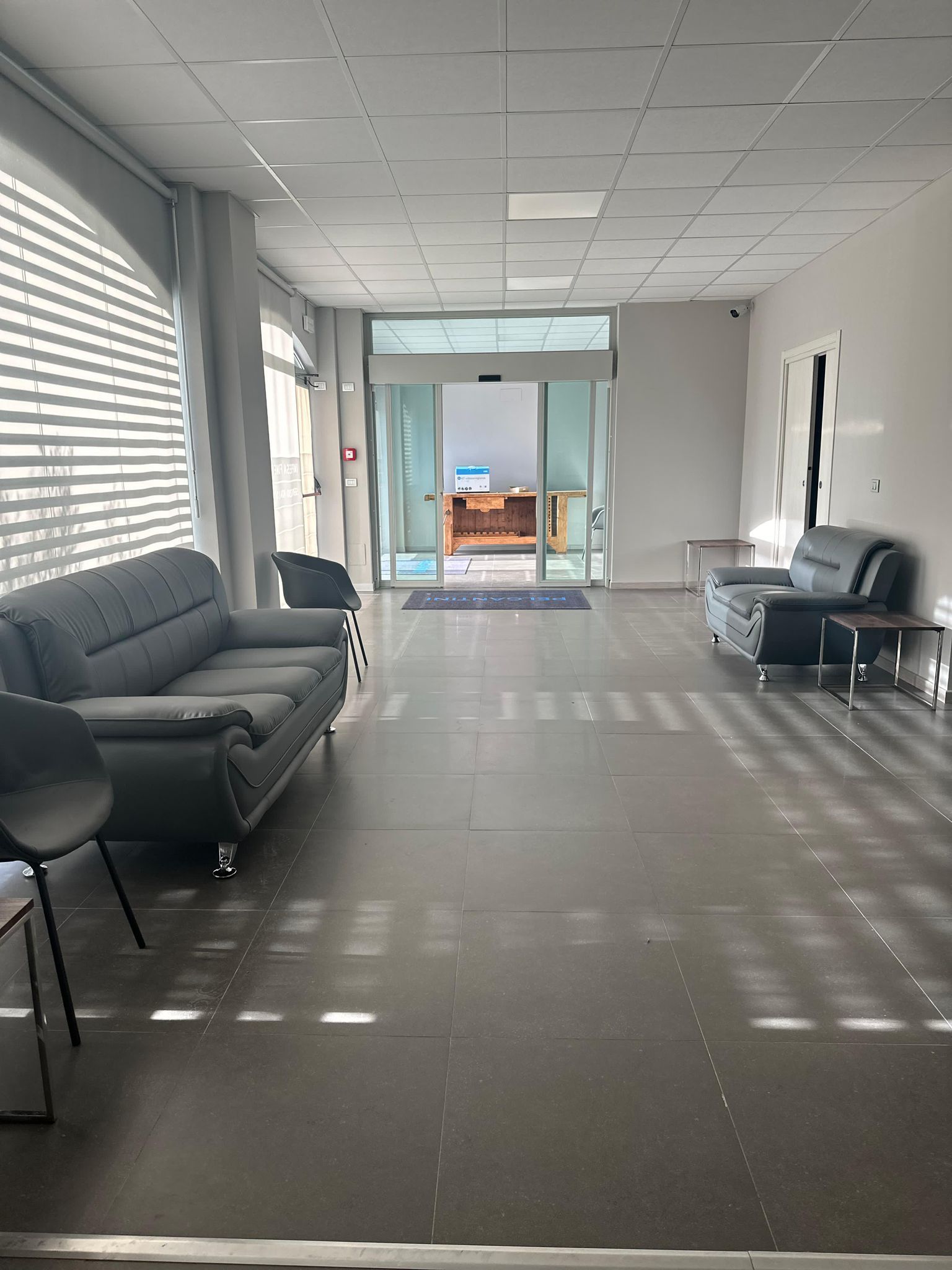 Sala d'attesa con divani e sedie grigie e una porta che dà sull'esterno. La luce del sole filtra attraverso le persiane.
