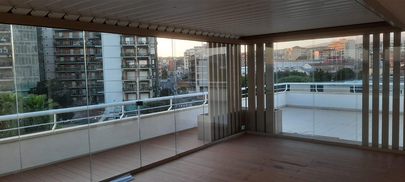 Un balcone coperto con pareti in vetro offre una vista sulla città. Supporti in legno incorniciano la vista e il pavimento è in legno.
