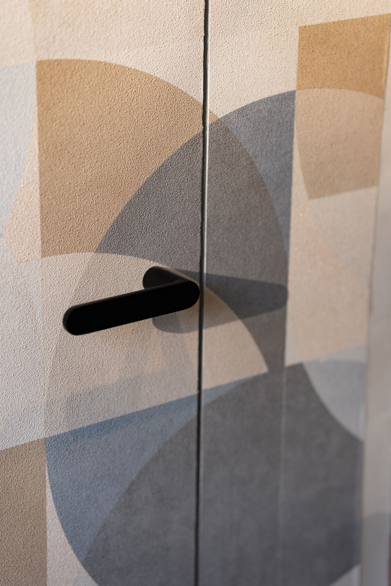 Maniglia nera su ante di armadi decorati con forme geometriche nei colori beige, blu e grigio.