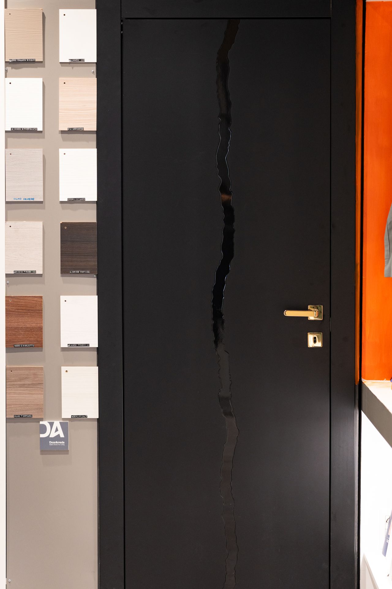 Porta nera con una crepa verticale frastagliata. Maniglia e serratura dorate. Campioni di legno a sinistra, parete arancione a destra.