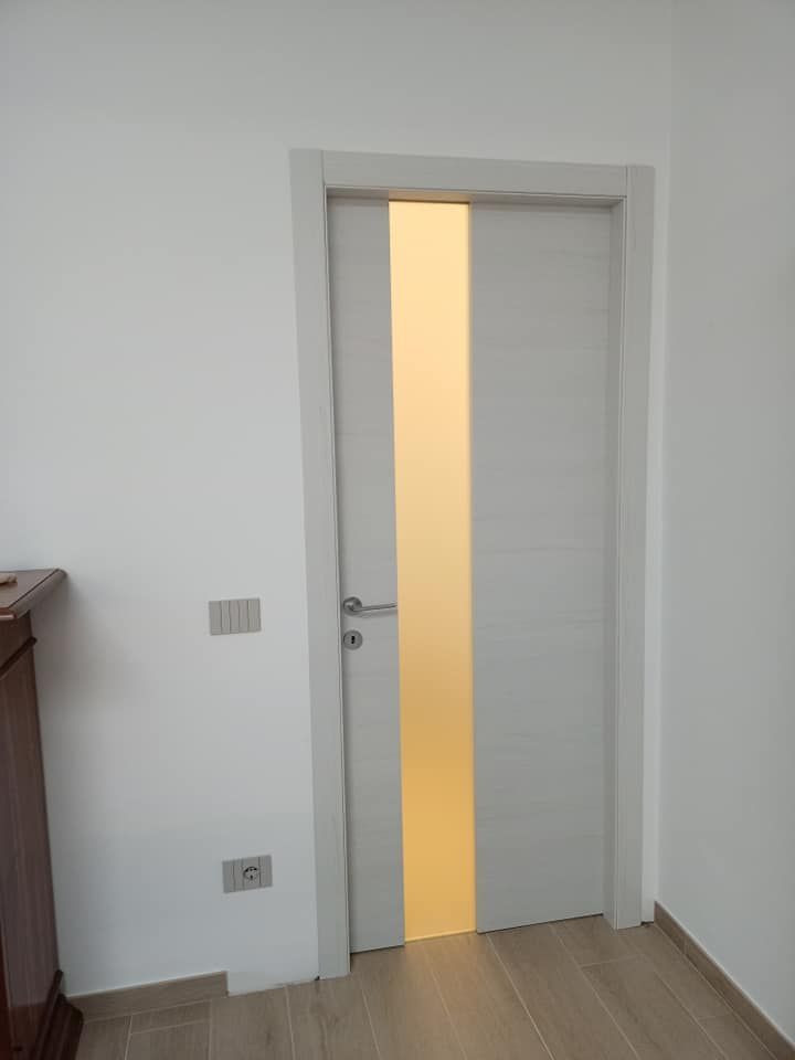 Porta grigia con pannello in vetro smerigliato, inserita in una parete bianca con pavimento di colore chiaro.