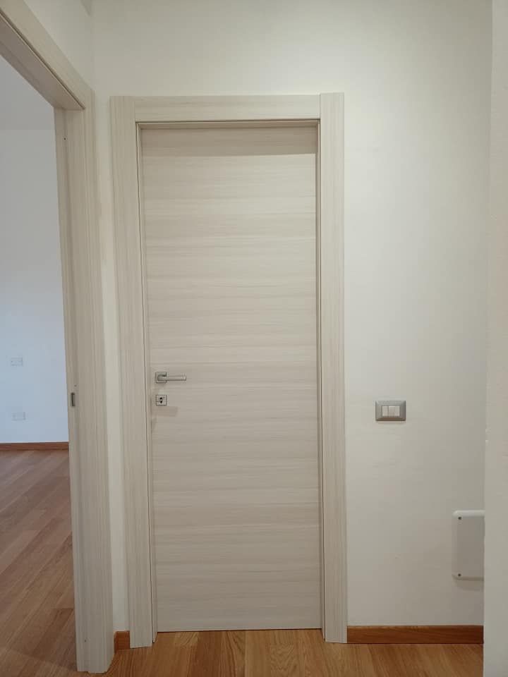 Porta interna bianca con telaio e maniglia coordinati, inserita in una parete bianca con pavimento in legno.