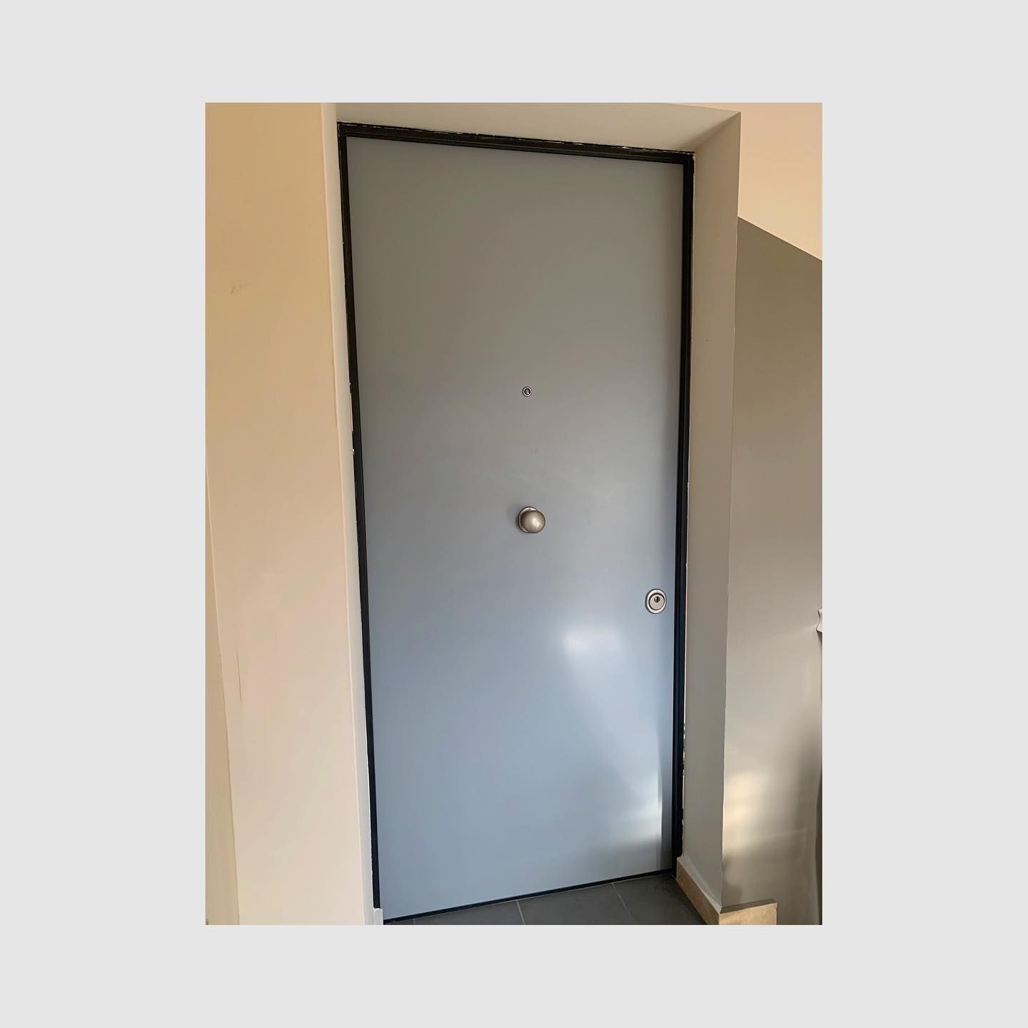 Porta grigia con finiture nere nel corridoio di un edificio, con una piccola maniglia e uno spioncino.