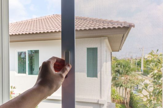 Mano che apre una zanzariera, con una casa e del verde visibili all'esterno.