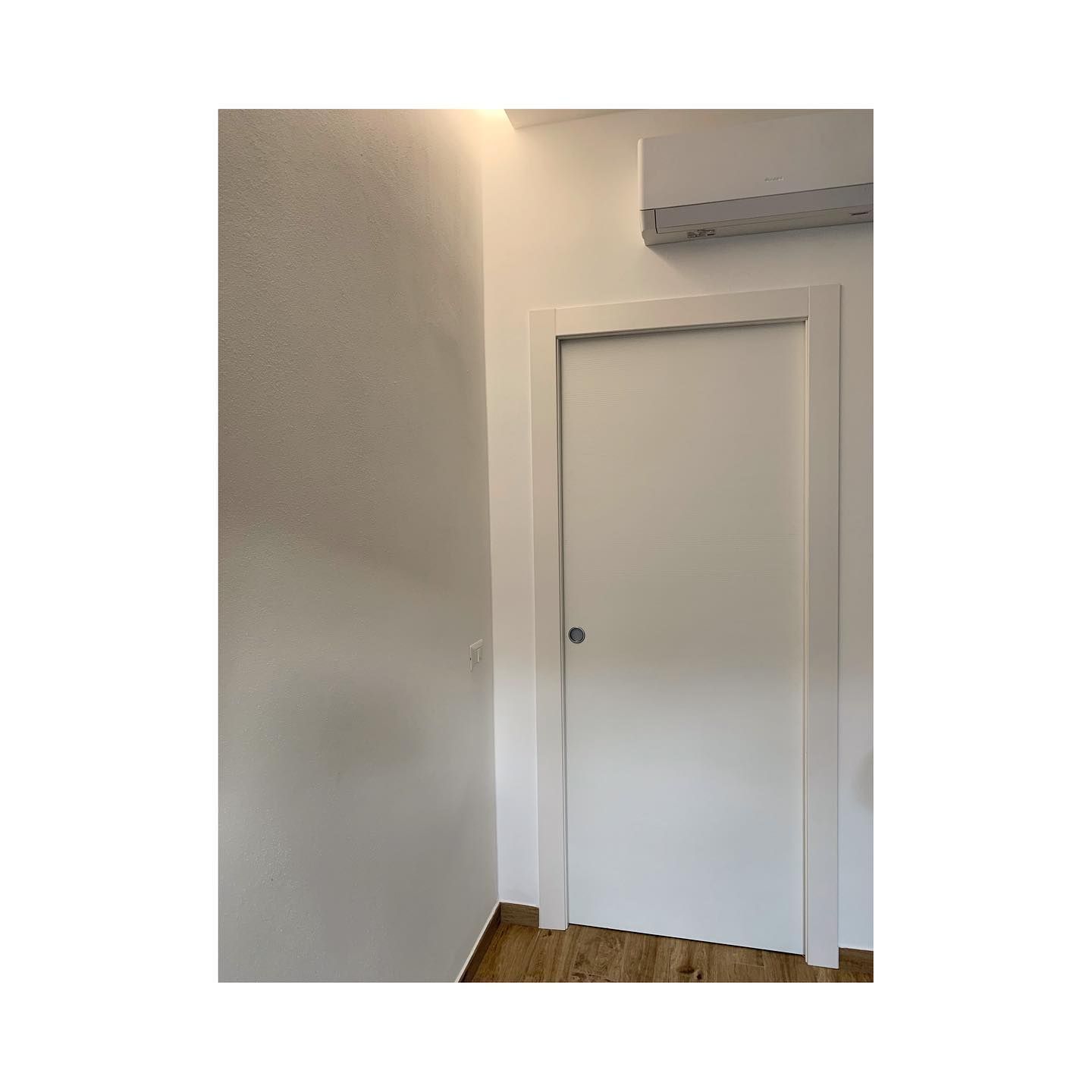 Porta bianca in una stanza con pareti bianche e pavimento in legno. Sopra la porta c'è un condizionatore.