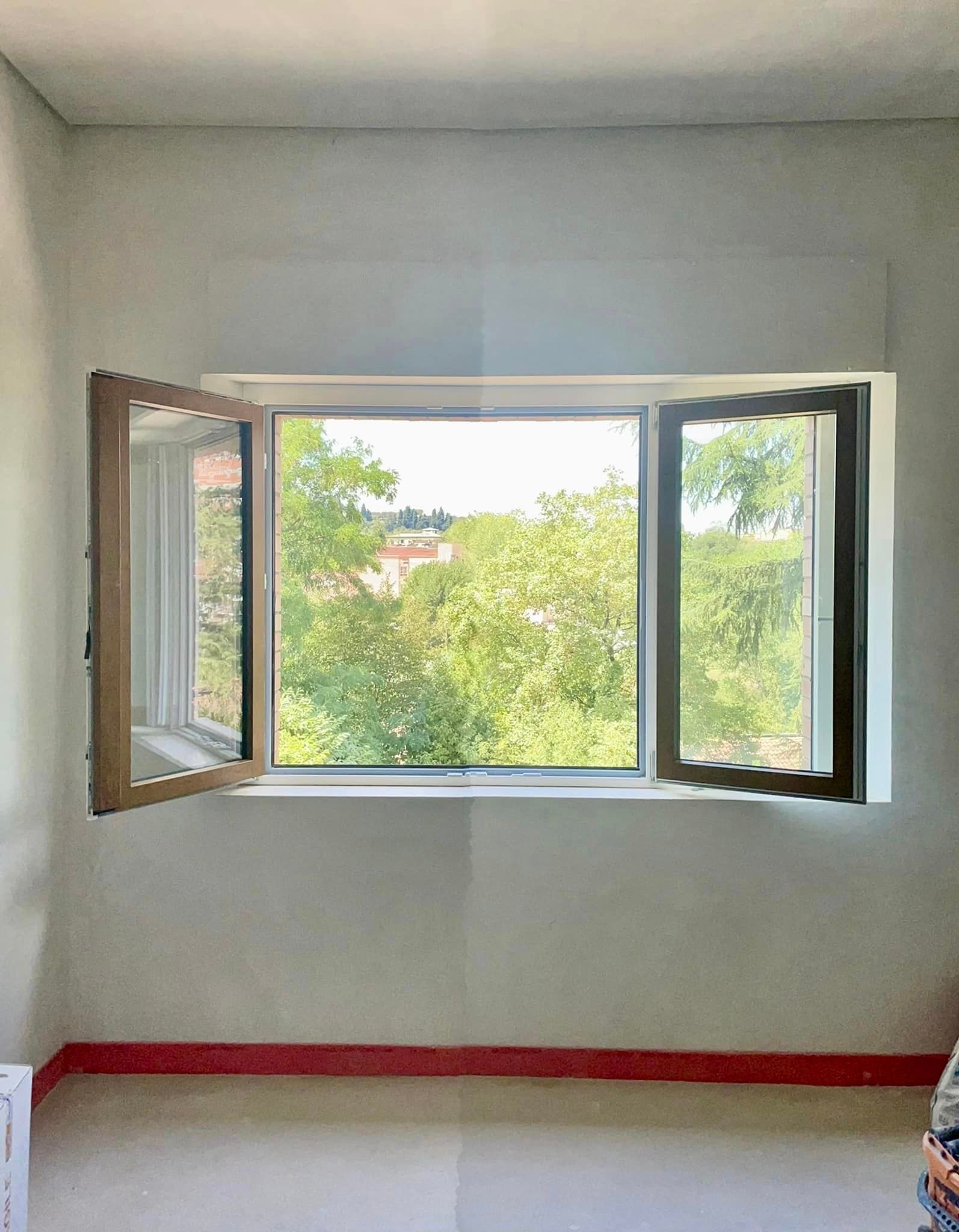 Finestra aperta con telaio in legno, con vista sugli alberi verdi. Pareti grigie, finiture rosse.
