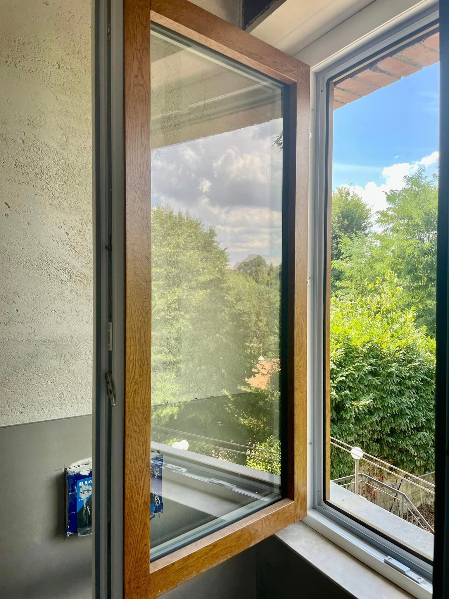 Finestra aperta con cornice marrone con vista sugli alberi e sul cielo. Parete interna e davanzale visibili.