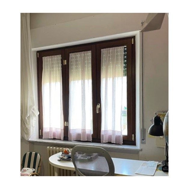 Finestra con cornice in legno, tende trasparenti, scrivania, sedia e termosifone.