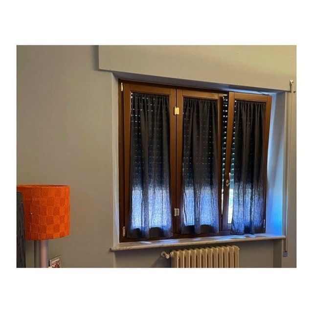 Finestra con cornice in legno e tende grigio scuro, un termosifone in basso e una lampada arancione a sinistra.