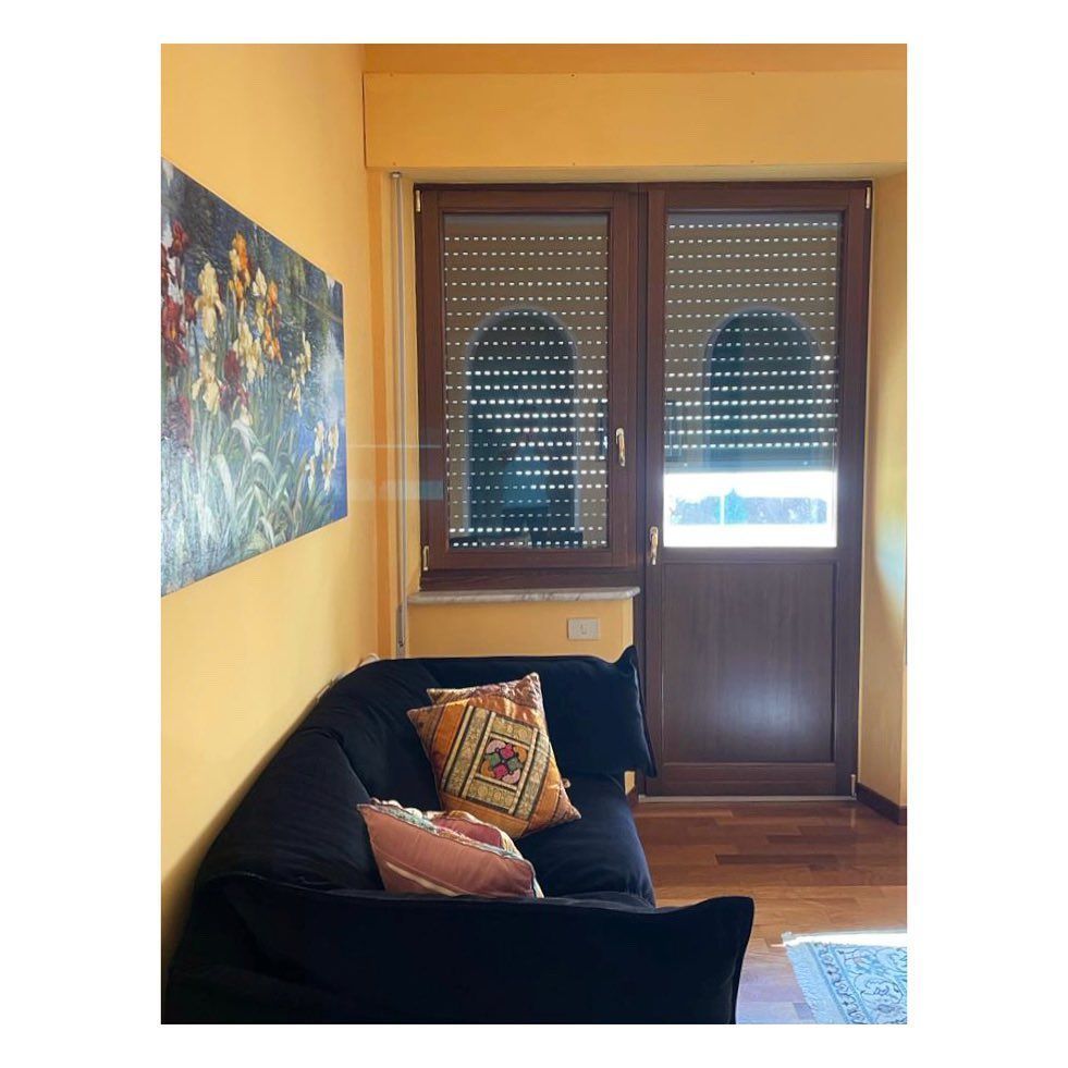 Divano nero in una stanza con una porta di legno e un dipinto colorato alla parete. Pareti gialle, persiane chiuse.
