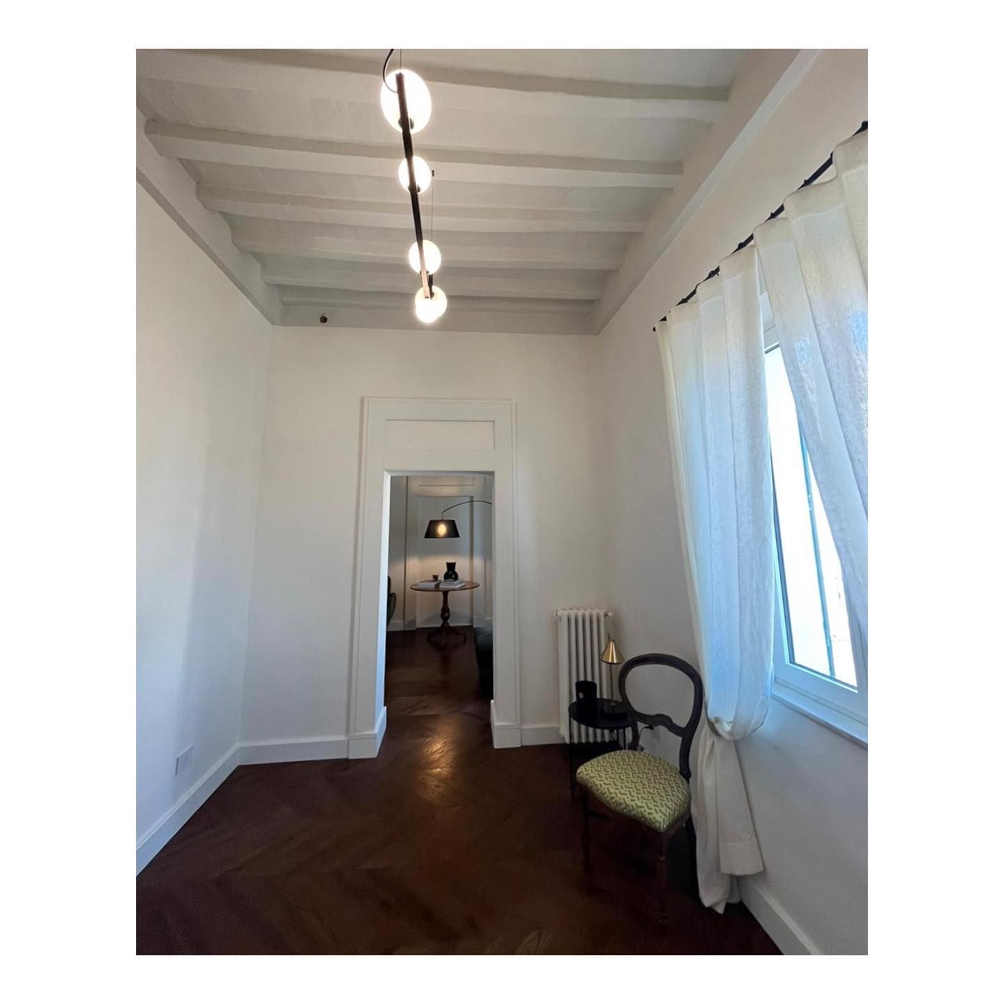 Stanza interna con pavimenti in legno, pareti bianche e una lampada sospesa. Una porta conduce a un'altra stanza.
