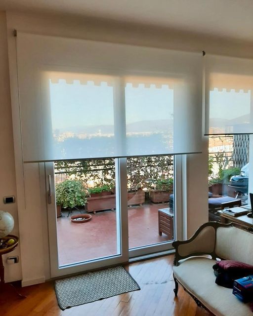 Le tende a rullo bianche coprono una porta scorrevole in vetro e le finestre che si affacciano su un balcone con piante.