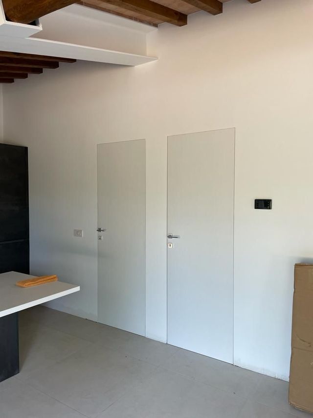 Due porte bianche a filo muro, con pavimento grigio e soffitto grezzo sopra.