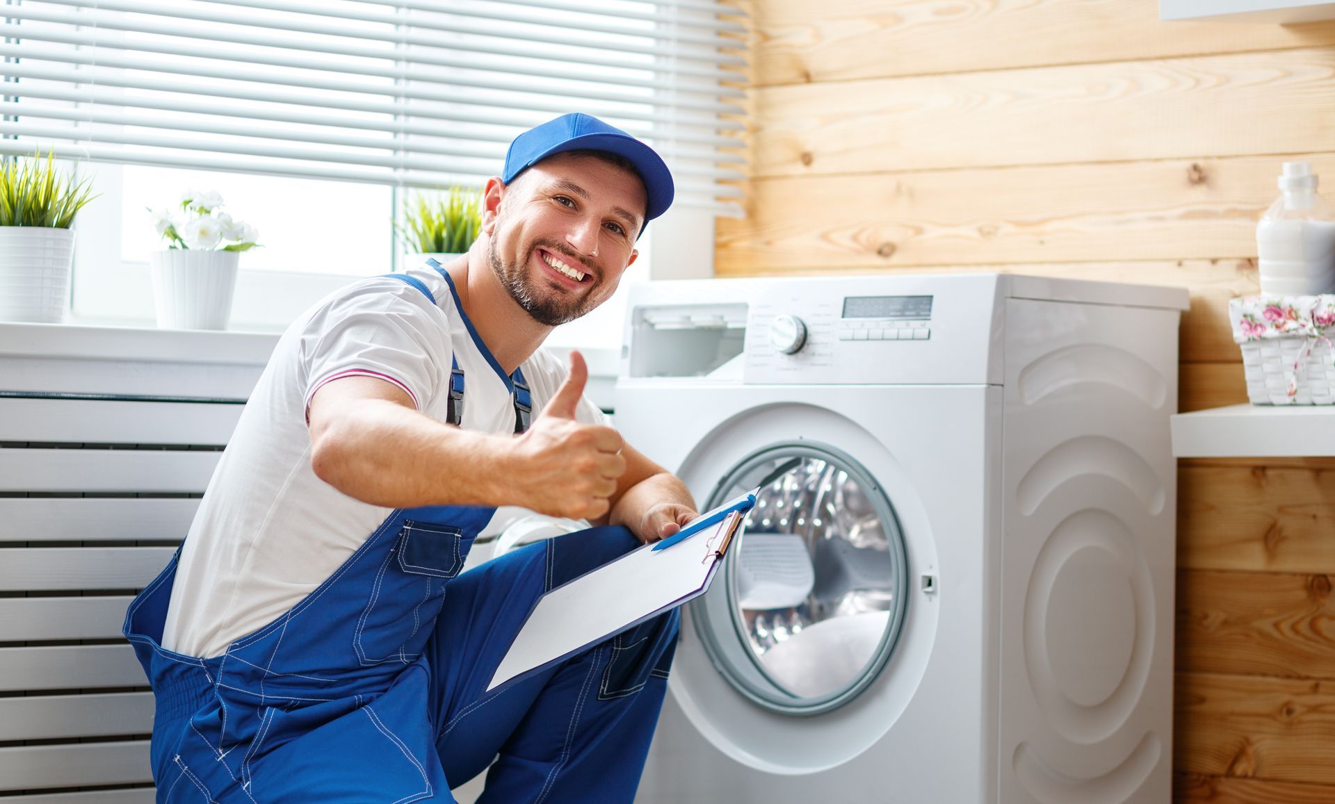 Smiling repairman by a washing machine, giving a thumbs up. Holding clipboard in front of laundry room window.