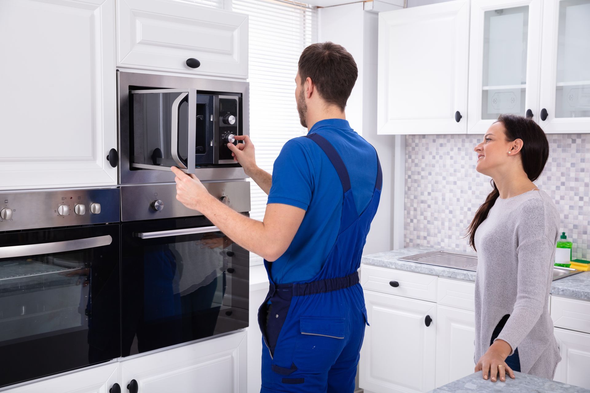A repairman in blue overalls fixing a built-in microwave, a woman watches in a white kitchen.