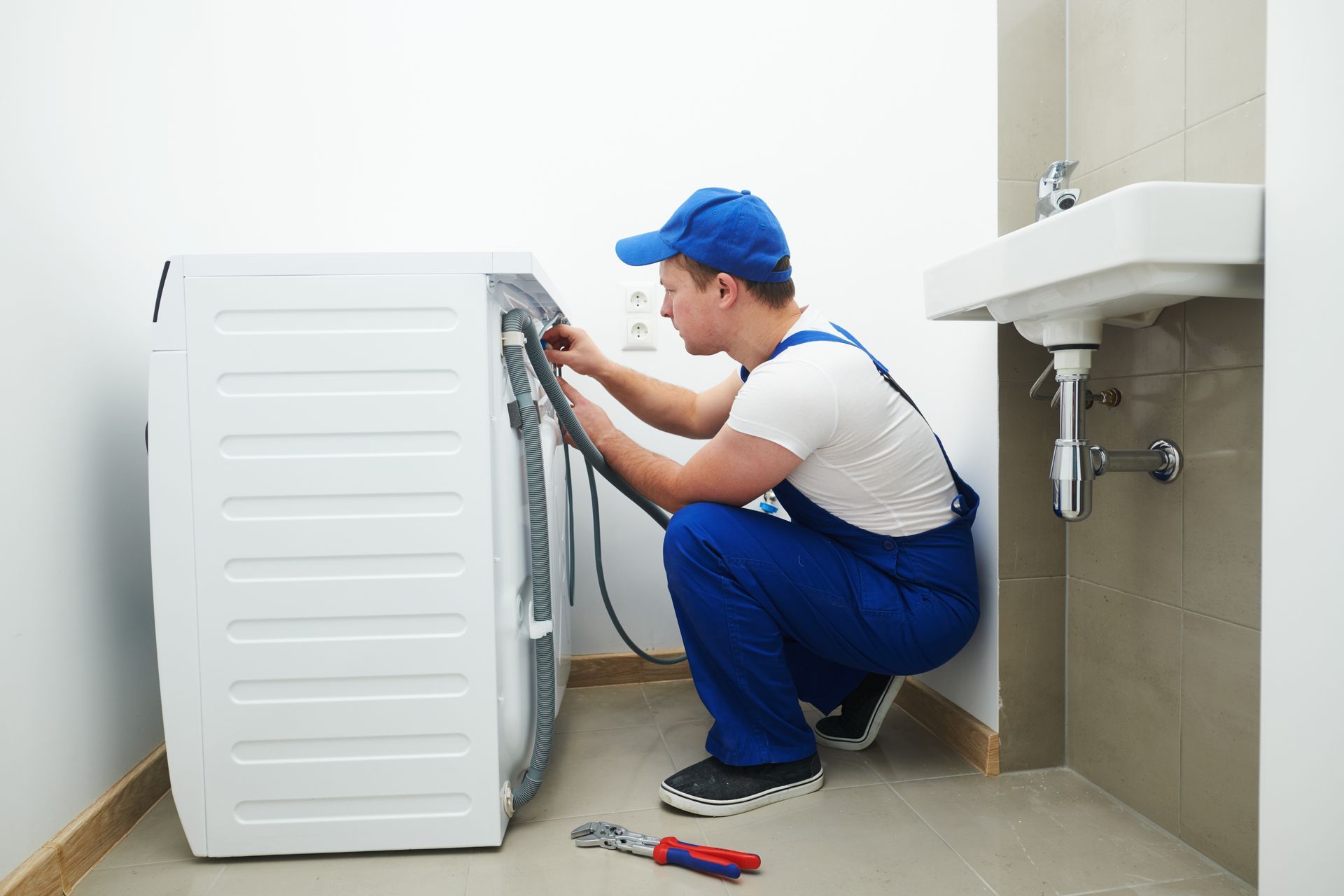 A man in blue overalls repairs a washing machine, focused on the internal components.