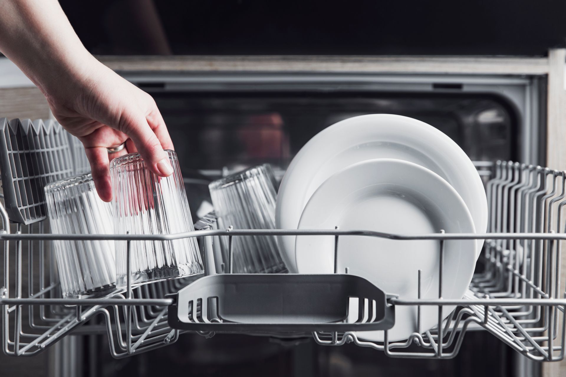 Hand placing a glass in a dishwasher rack filled with plates and glasses.