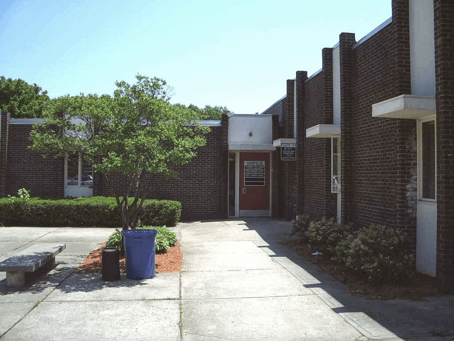 Brick building entrance with a sidewalk, trees, and a blue planter in the foreground