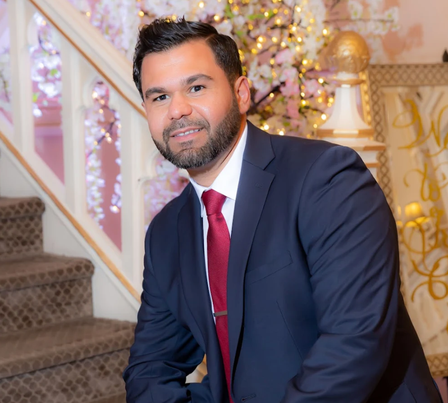 Man in navy suit and red tie smiling on a staircase with soft decorative lights and gold accents