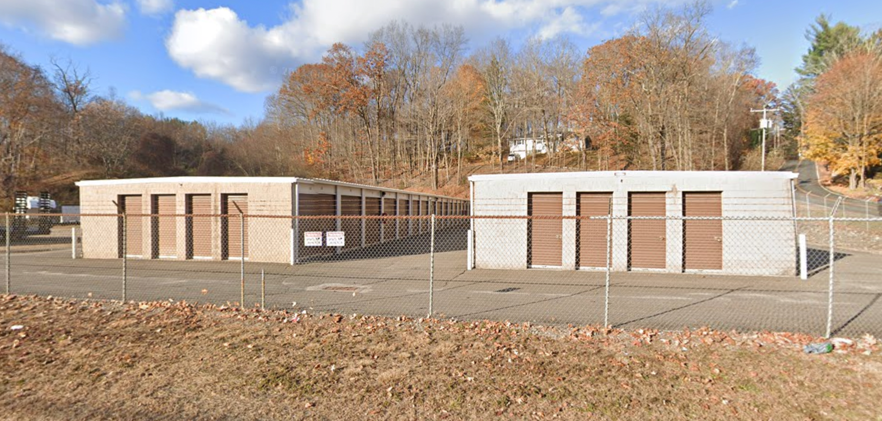 Two small utility buildings behind a chain-link fence in a grassy field with autumn trees and a blue sky