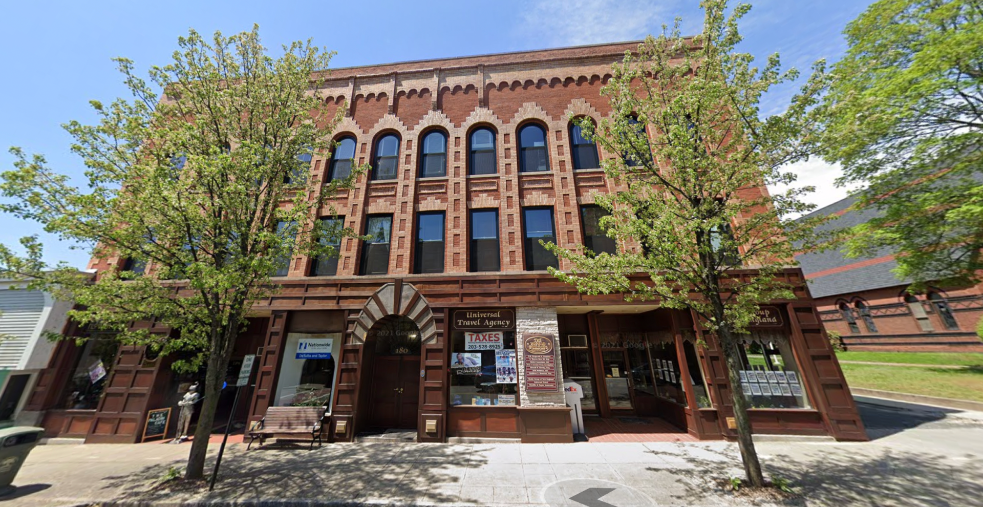 Historic red-brick building with arched windows and storefronts, framed by trees on a sunny street.