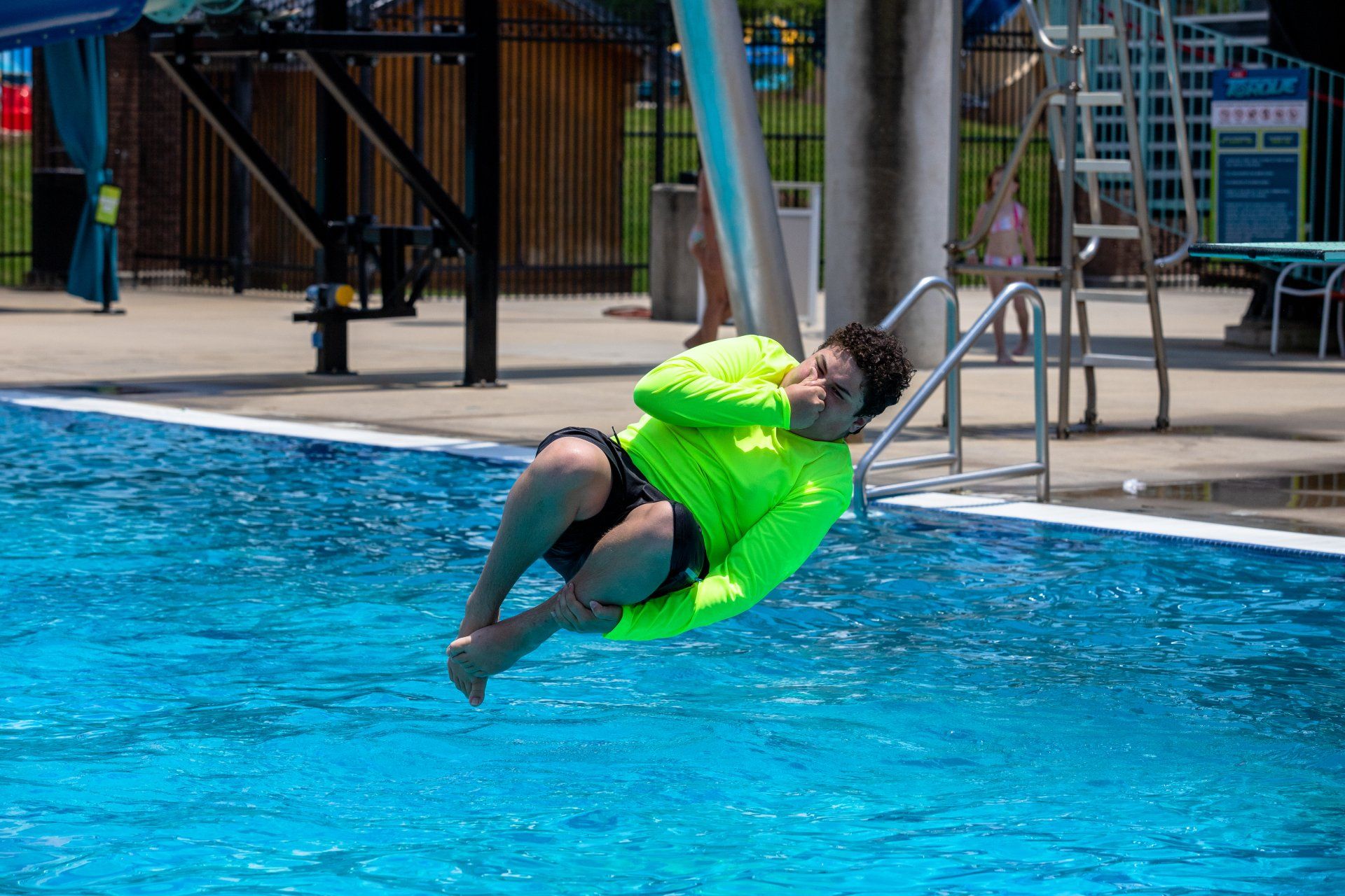 Person in a neon green shirt splashes into a pool near the edge, with a ladder and deck in the background.