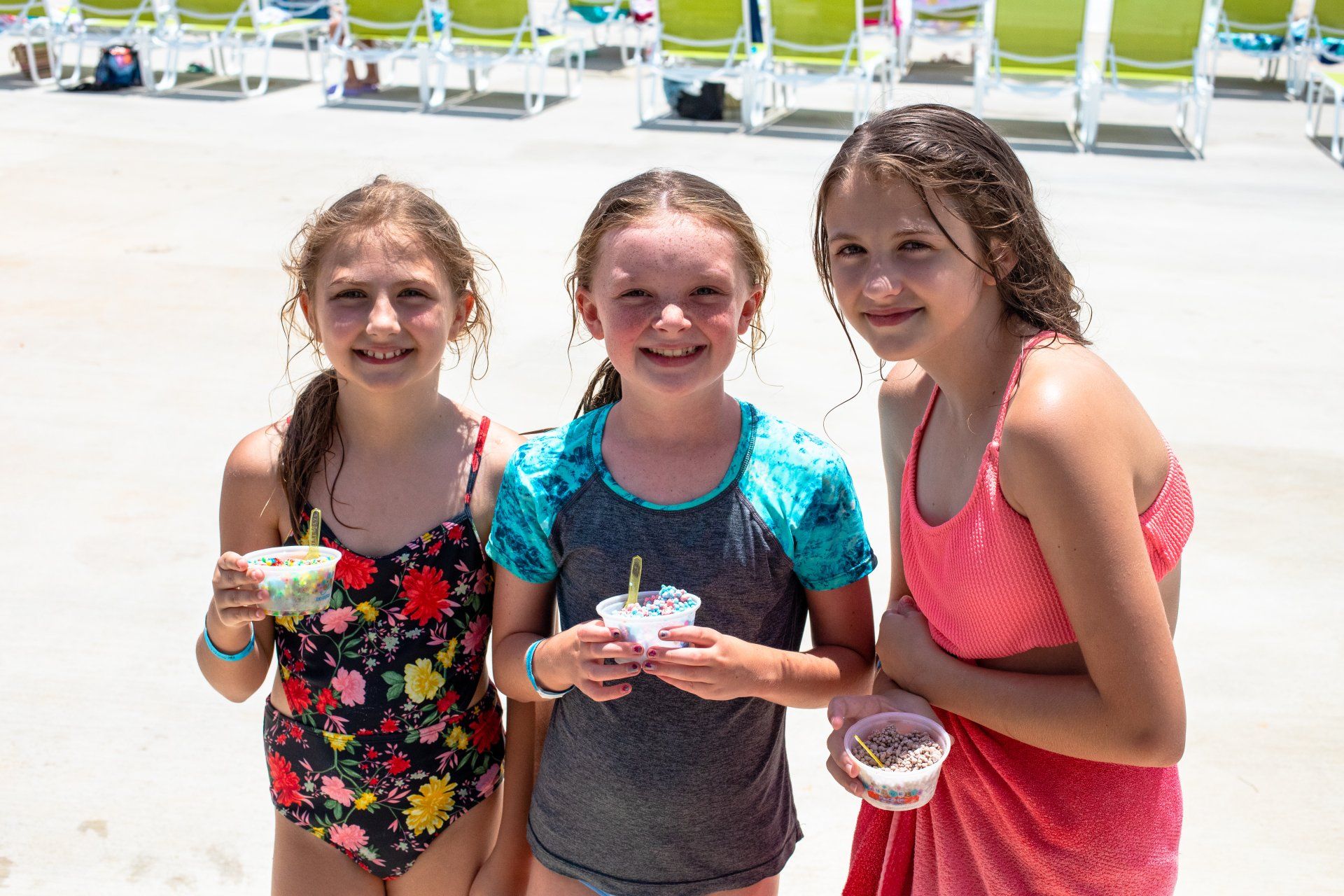 Three smiling kids in swimsuits holding drinks by a pool on a sunny day