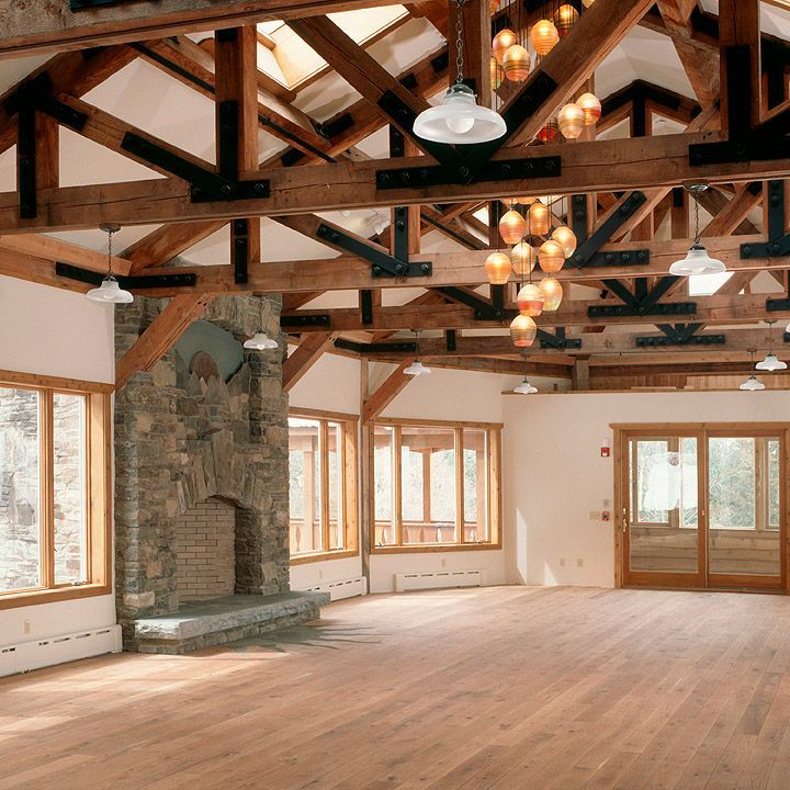 Spacious rustic hall with exposed beams, stone fireplace, wood floors, and large windows under hanging lights