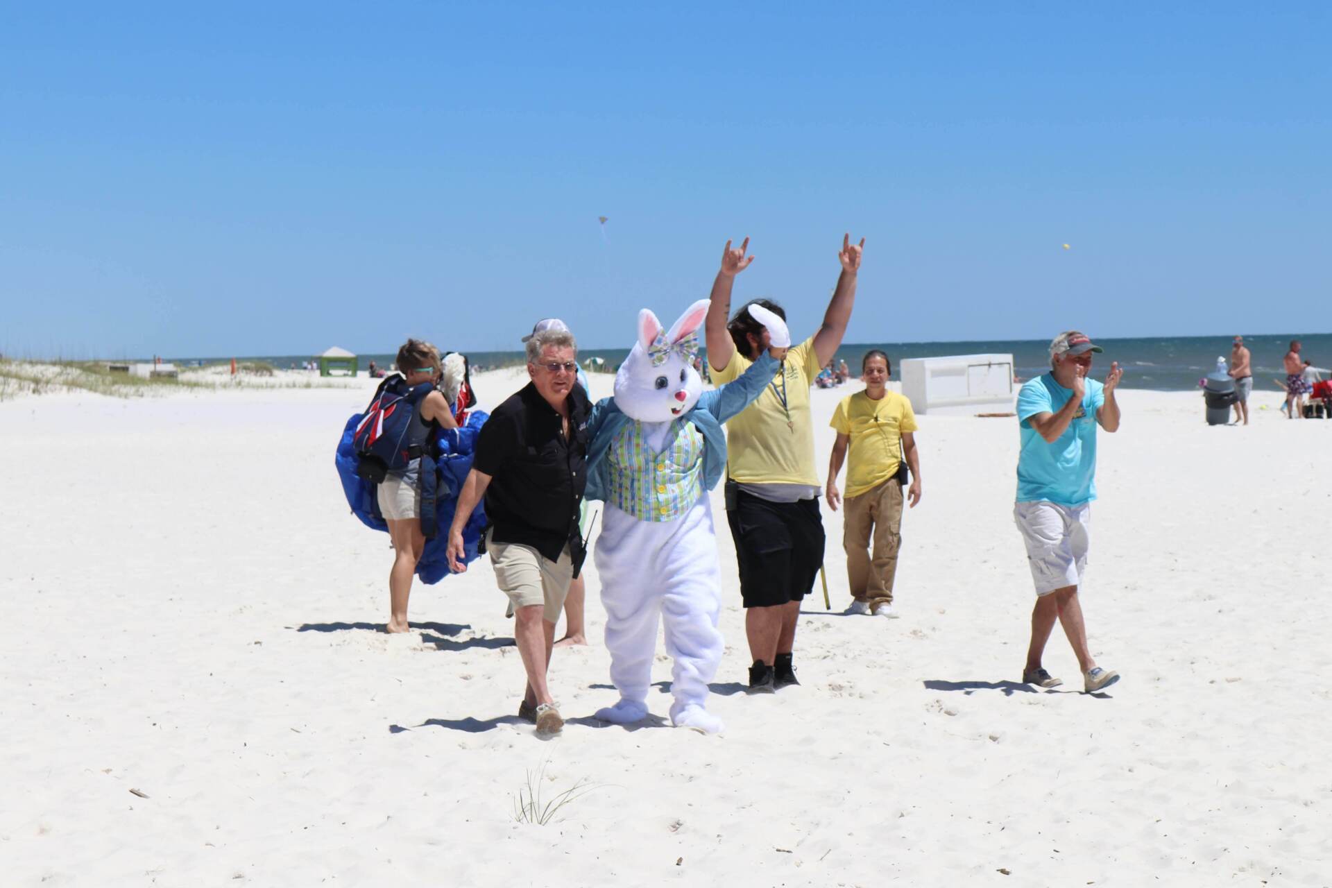 A costumed Easter bunny walks along a sunny beach with a group of people, with one person cheering and another behind.